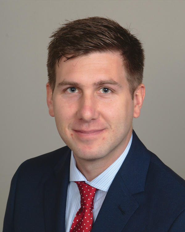 A man with short brown hair wearing a blue suit, light blue shirt, and red polka dot tie, poses in front of a plain light background, smiling slightly at the camera.