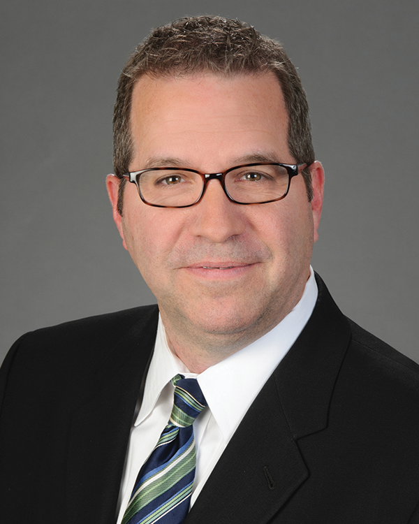 A man wearing glasses, a dark suit, a white shirt, and a green and blue striped tie poses for a professional headshot against a plain gray background.