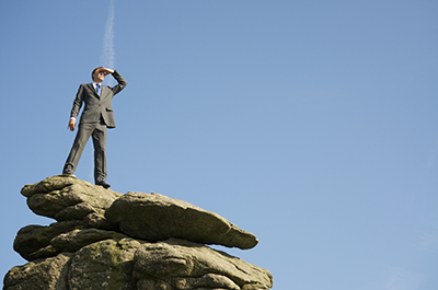 A man in a suit stands on top of a rocky outcrop, shading his eyes as he looks into the distance.