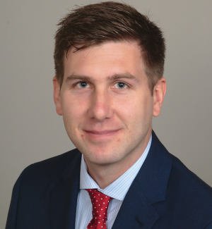 A man with short brown hair wearing a blue suit, light blue shirt, and red polka dot tie, poses in front of a plain light background, smiling slightly at the camera.