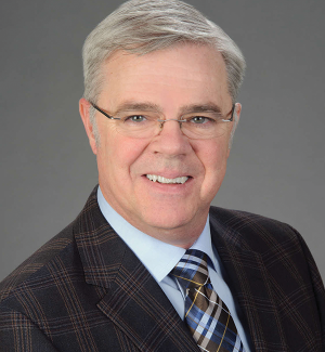A smiling older man with gray hair and glasses wears a plaid suit jacket, blue shirt, and plaid tie. The background is plain and gray.
