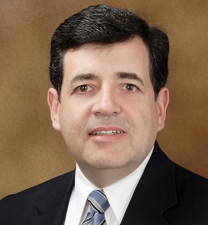 A man in a dark suit, striped tie, and white shirt poses against a brown background, looking forward with a neutral expression.