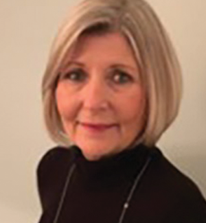 A woman with short, blonde hair is wearing a black top and necklace, standing in front of a plain light background, looking at the camera with a slight smile.