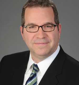 A man wearing glasses, a dark suit, a white shirt, and a green and blue striped tie poses for a professional headshot against a plain gray background.