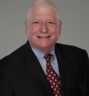 A bald man wearing a black suit, light blue shirt, and red patterned tie, smiling at the camera against a plain gray background.