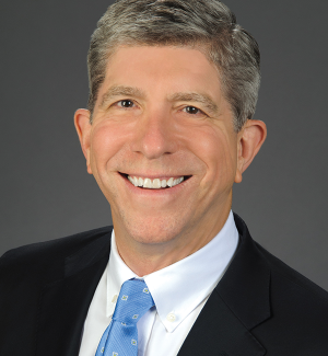 Smiling man with short gray hair wearing a dark suit, white shirt, and light blue tie, posed in front of a plain gray background.