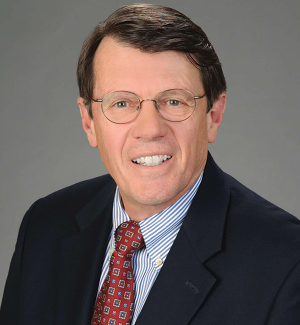A man wearing glasses, a dark suit, a white and blue striped shirt, and a red patterned tie, smiling against a plain gray background.