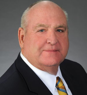 A middle-aged man with a bald head, wearing a dark suit jacket, white shirt, and yellow striped tie, poses in front of a plain gray background.