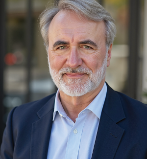 A middle-aged man with gray hair and a beard, wearing a navy suit and light blue shirt, stands outside in front of glass doors, smiling gently at the camera.