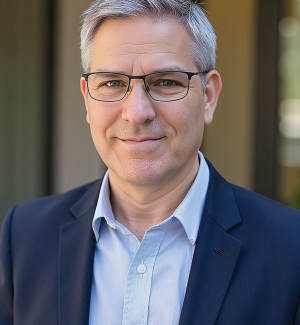 A middle-aged man with short gray hair and glasses, wearing a dark blazer over a light blue shirt, smiles at the camera in an outdoor setting with blurred background.